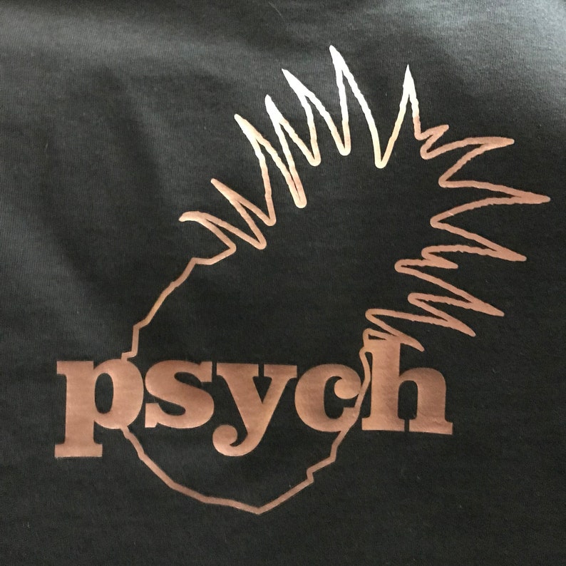 May include: A black t-shirt featuring a metallic copper-colored graphic of a pineapple silhouette. The word "psych" is printed in lowercase, bold, copper-colored letters across the lower portion of the pineapple design on the front of the shirt.