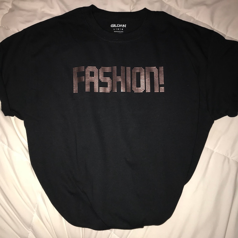 May include: A black short-sleeved t-shirt with a crew neckline. The center of the chest features the word FASHION! printed in a metallic copper-colored dot matrix pattern. The interior collar tag displays the Gildan brand name and size L/G/G.
