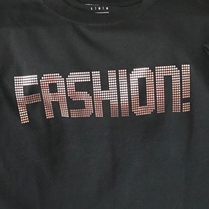 May include: A black t-shirt featuring the word FASHION! printed in a dot-matrix style graphic. The letters are composed of small, individual circles that create a gradient effect, transitioning from a light metallic copper color to a darker bronze tone.