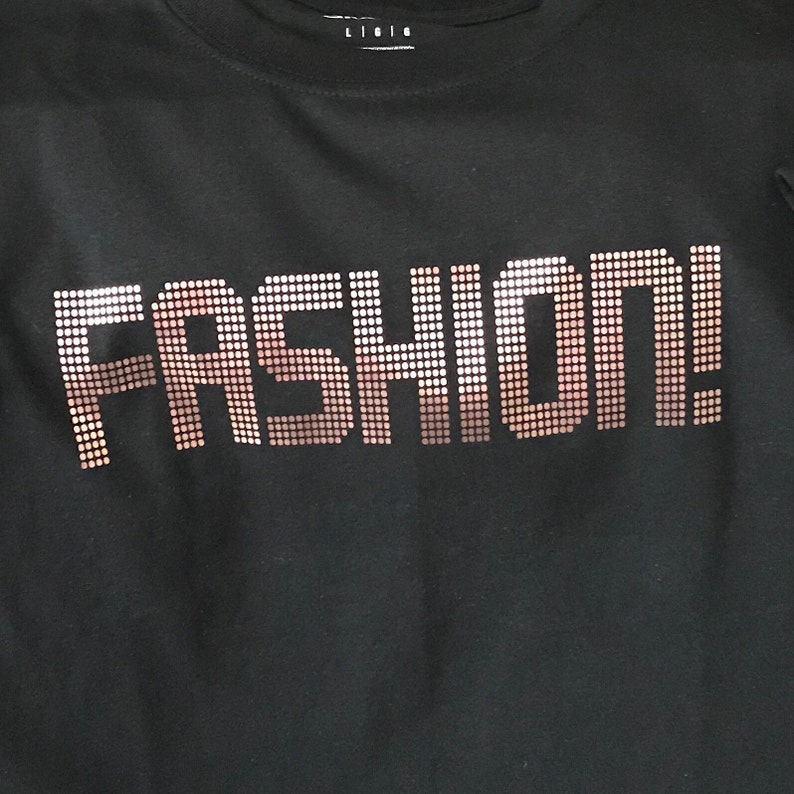 May include: A black t-shirt featuring the word FASHION! printed in a dot-matrix style graphic. The letters are composed of small, individual circles that create a gradient effect, transitioning from a light metallic copper color to a darker bronze tone.