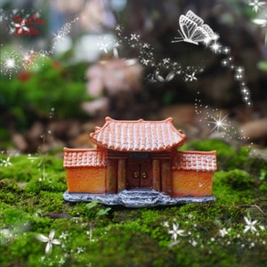 Op de afbeelding: Een miniatuur oranje en bruine tempel met een betegeld dak en een dubbele deuropening. De tempel is omgeven door groen mos en witte bloemen.