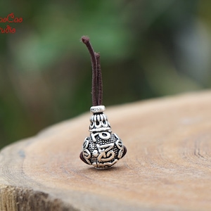 May include: A silver pendant with detailed patterns and a dark brown cord. The pendant has a bulbous shape with detailed carvings and a small loop at the top. The cord is attached to the pendant with a small silver ring.