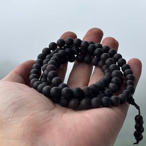 Puede incluir: Un collar de cuentas de madera marrón oscuro, probablemente un mala de oración, sostenido en una mano abierta. Las cuentas redondas y mate tienen una borla anudada. El fondo es suave, gris claro.