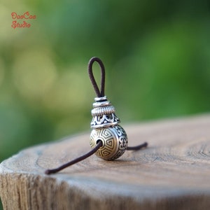 Puede incluir: Una cuenta de plata y oro con diseños intrincados, colgando de un cordón marrón. La cuenta tiene una pequeña tapa de plata en la parte superior.