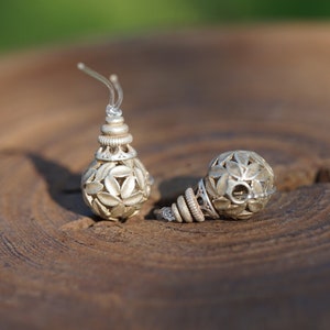 Könnte beinhalten: Ein Paar silberfarbene Ohrringe mit floralem Design. Ein Ohrring steht aufrecht und zeigt den Ohrhaken, während der andere auf der Seite liegt. Die Ohrringe haben ein detailliertes, durchbrochenes Design und liegen auf einer Holzoberfläche.