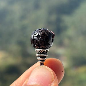 May include: A dark, intricately carved bead with a spherical shape. The bead features a detailed pattern and is attached to a silver-coloured metal base. The bead is held by a person's fingers against a blurred green background.