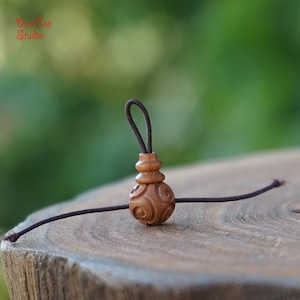 May include: A brown wooden bead with a carved pattern, attached to a brown elastic cord. The bead has a smaller, stacked bead on top.