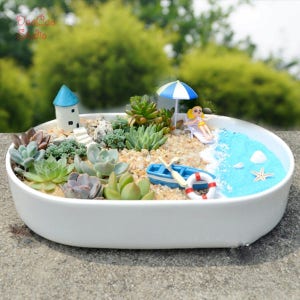 May include: A white oval planter featuring a miniature beach scene. The scene includes a small blue house, a white sandy beach, a blue ocean, a small boat, a life preserver, a starfish, a seashell, and a miniature person sitting on a beach chair under a blue and white umbrella.