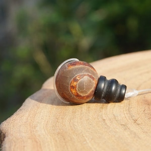 Peut inclure: Une perle d'agate brune avec un motif en spirale blanc et orange, enfilée sur un cordon élastique transparent avec une perle noire à l'extrémité.
