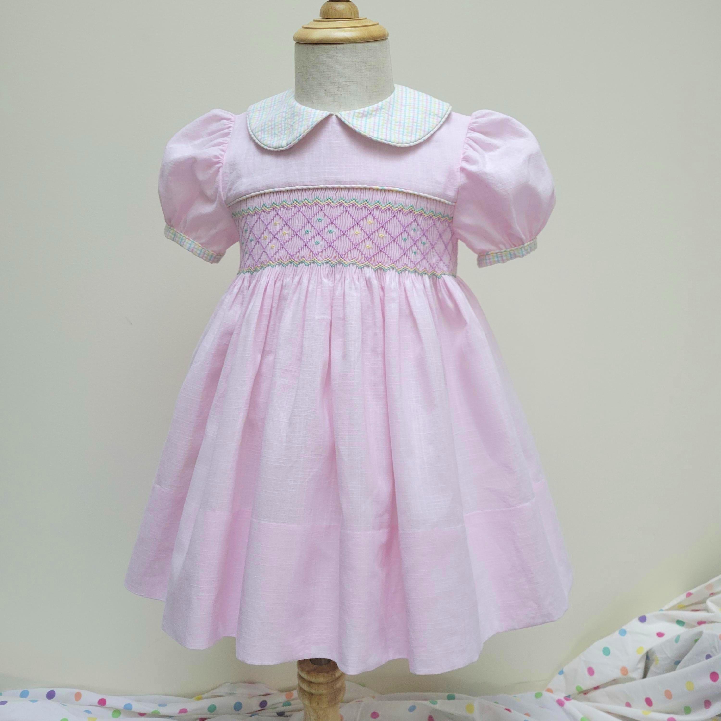 May include: A light pink dress featuring short puff sleeves, a Peter Pan collar with a light blue and white gingham pattern, and a smocked bodice with intricate diamond-shaped embroidery in shades of pink, blue, and yellow on the chest.