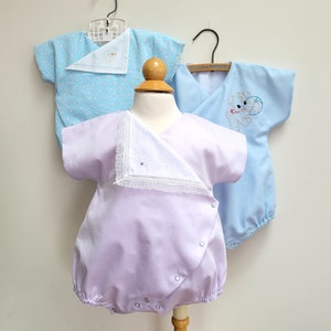 Puede incluir: Tres bodies de bebé, uno azul claro con un diseño bordado blanco, uno azul claro con un diseño bordado blanco y uno lavanda con un borde de encaje blanco y un diseño bordado blanco.