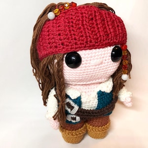 Pode incluir: Boneca de crochê com cabelo castanho, um chapéu vermelho e um colete azul e branco. A boneca está usando botas marrons e tem grandes olhos pretos.