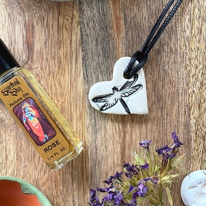May include: A white ceramic heart pendant with a dragonfly design hangs from a black cord. The pendant is on a wooden surface with a small bottle of rose perfume, dried flowers, and a white ceramic dragonfly.