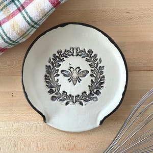 Op de afbeelding: Een witte keramische lepelsteun met een zwarte rand, met een bij en laurierkrans ontwerp. Een geruite keukendoek en een garde zijn ook zichtbaar op een houten oppervlak. Keukengerei.