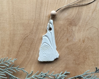 May include: A white ceramic ornament shaped like the state of New Hampshire, with a spiral design. It hangs from a natural twine cord and a wooden bead. The ornament is displayed on a wooden surface with a sprig of greenery.