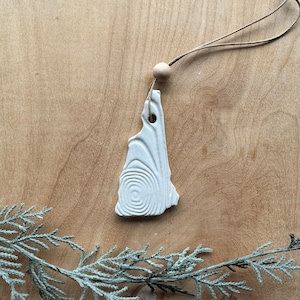 May include: A white ceramic ornament shaped like the state of New Hampshire, with a spiral design. It hangs from a natural twine cord and a wooden bead. The ornament is displayed on a wooden surface with a sprig of greenery.