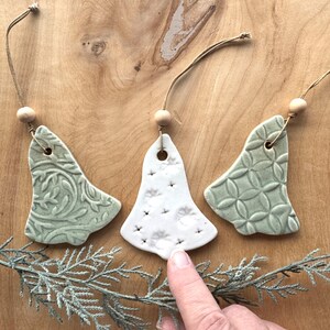 May include: Three ceramic Christmas tree ornaments in the shape of bells. The ornaments are white with green accents and have different patterns. One ornament has a white background with green dots, another has a green background with a white floral pattern, and the third has a green background with a white geometric pattern.