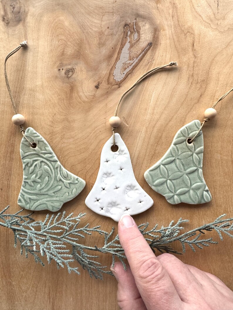 May include: Three ceramic Christmas tree ornaments in the shape of bells. The ornaments are white with green accents and have different patterns. One ornament has a white background with green dots, another has a green background with a white floral pattern, and the third has a green background with a white geometric pattern.