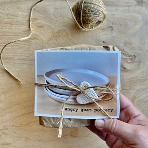 May include: A package wrapped in brown paper with twine and a white clay heart. The package contains a set of white ceramic plates. The package is labeled "angry goat pottery".