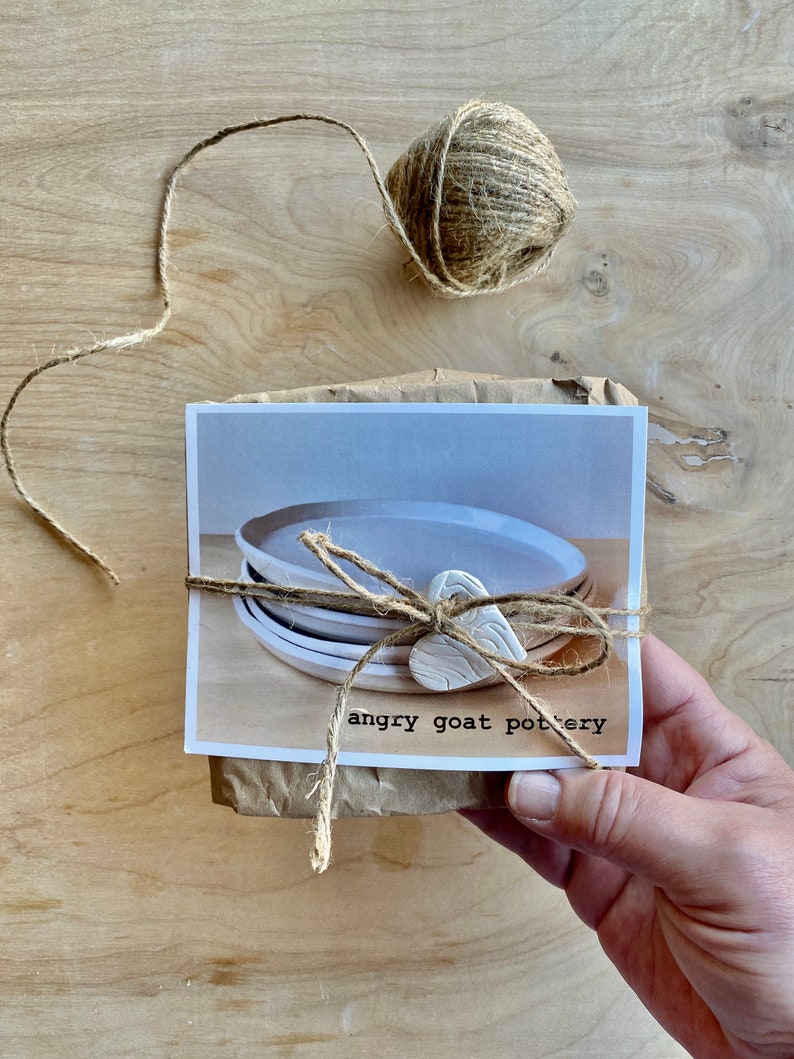 May include: A package wrapped in brown paper with twine and a white clay heart. The package contains a set of white ceramic plates. The package is labeled "angry goat pottery".