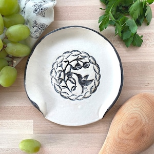 May include: A white ceramic spoon rest with a black rim and a central design of a hummingbird and foliage. The spoon rest has two small side indents and is surrounded by a wooden spoon, grapes, and parsley.