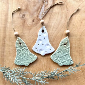 May include: Three ceramic Christmas tree ornaments in the shape of bells. Two are green with different embossed patterns and one is white with a star pattern.