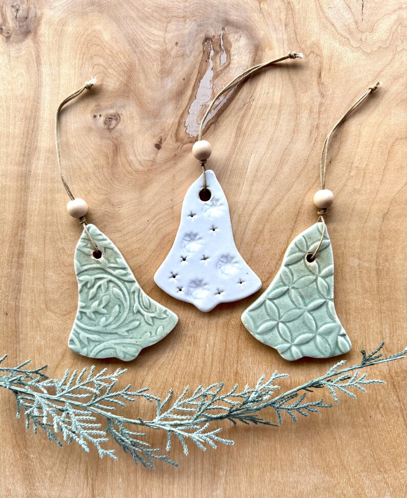 May include: Three ceramic Christmas tree ornaments in the shape of bells. Two are green with different embossed patterns and one is white with a star pattern.