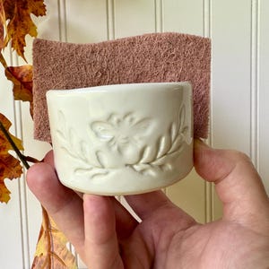 May include: A cream-colored ceramic sponge holder with a debossed bee and leaf design on the front. The holder contains a rectangular, textured, dusty rose-colored scouring sponge that extends above the top rim of the ceramic container.