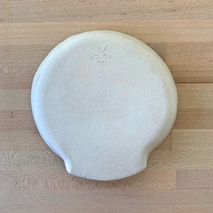 Op de afbeelding: Een witte keramische plaat met een textuur en een unieke vorm. De plaat heeft een klein, decoratief ontwerp dat in de buurt van de bovenkant is ge&euml;tst. De plaat staat op een lichtgekleurd houten oppervlak.