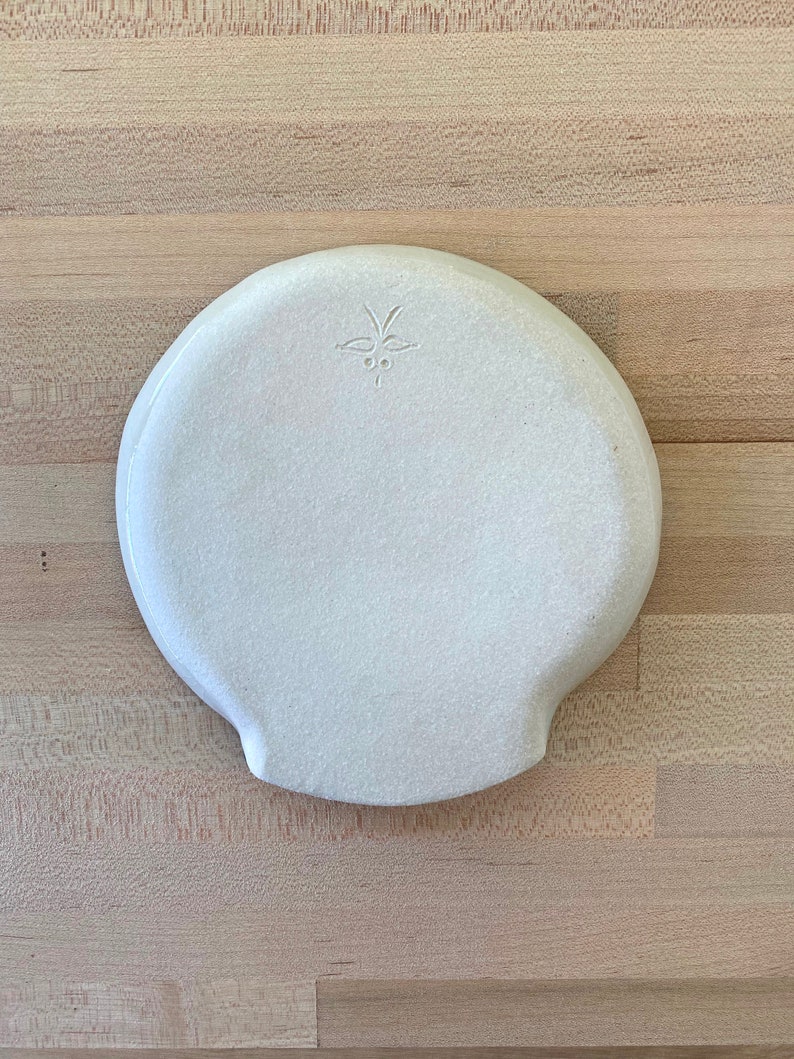 Op de afbeelding: Een witte keramische plaat met een textuur en een unieke vorm. De plaat heeft een klein, decoratief ontwerp dat in de buurt van de bovenkant is ge&euml;tst. De plaat staat op een lichtgekleurd houten oppervlak.