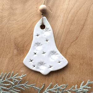 May include: White ceramic bell ornament with a textured surface and a hole at the top for hanging. The bell is decorated with a pattern of small circles and stars.