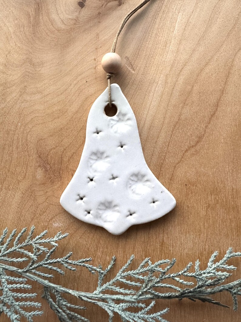 May include: White ceramic bell ornament with a textured surface and a hole at the top for hanging. The bell is decorated with a pattern of small circles and stars.