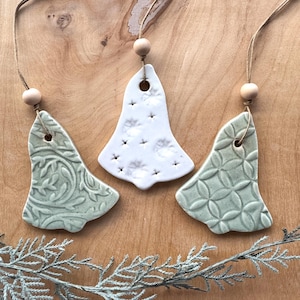 May include: Three ceramic bell ornaments with a rustic, farmhouse style. Two are green with a floral pattern and one is white with a dotted pattern. Each ornament has a hole at the top for hanging and a wooden bead on a twine string.