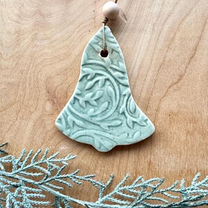 May include: A light green ceramic bell ornament with a floral pattern. The bell has a hole at the top for hanging and is attached to a string with a wooden bead.
