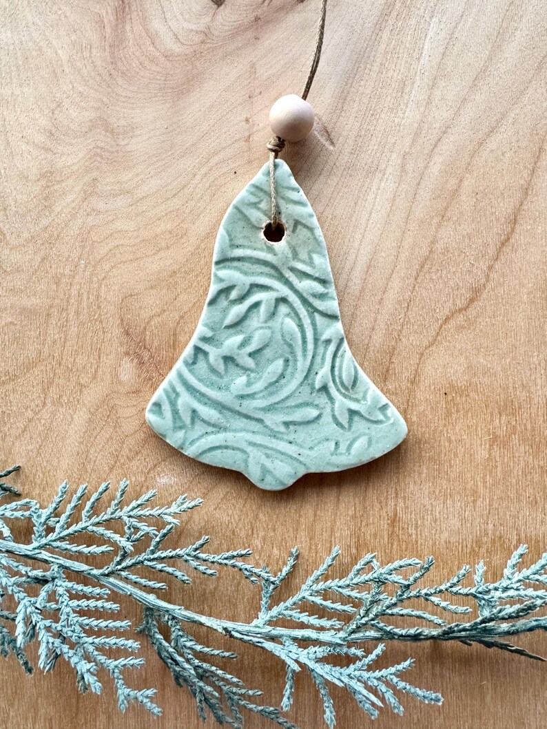 May include: A light green ceramic bell ornament with a floral pattern. The bell has a hole at the top for hanging and is attached to a string with a wooden bead.