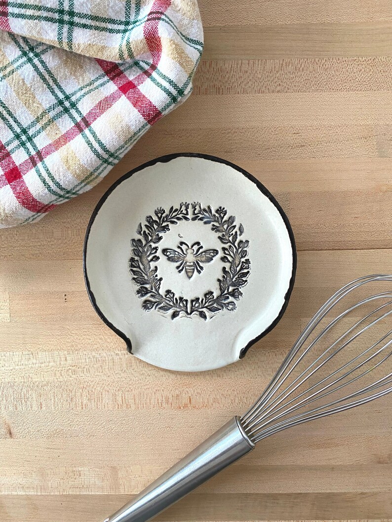 Op de afbeelding: Een witte keramische lepelsteun met een zwarte rand, met een bij en laurierkrans ontwerp. Een geruite keukendoek en een garde zijn ook zichtbaar op een houten oppervlak. Keukengerei.