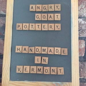 以下が含まれることがあります： 「Angry Goat Pottery Handmade in Vermont」と書いた文字タイルが置かれた黒板です。