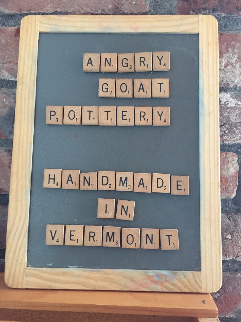 以下が含まれることがあります： 「Angry Goat Pottery Handmade in Vermont」と書いた文字タイルが置かれた黒板です。
