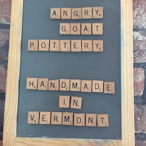 Op de afbeelding: Een ingelijst schoolbord met de woorden "ANGRY GOAT POTTERY HANDMADE IN VERMONT" geschreven met houten lettertegels. De lijst is van licht hout en de achtergrond is een donkergrijs schoolbord.
