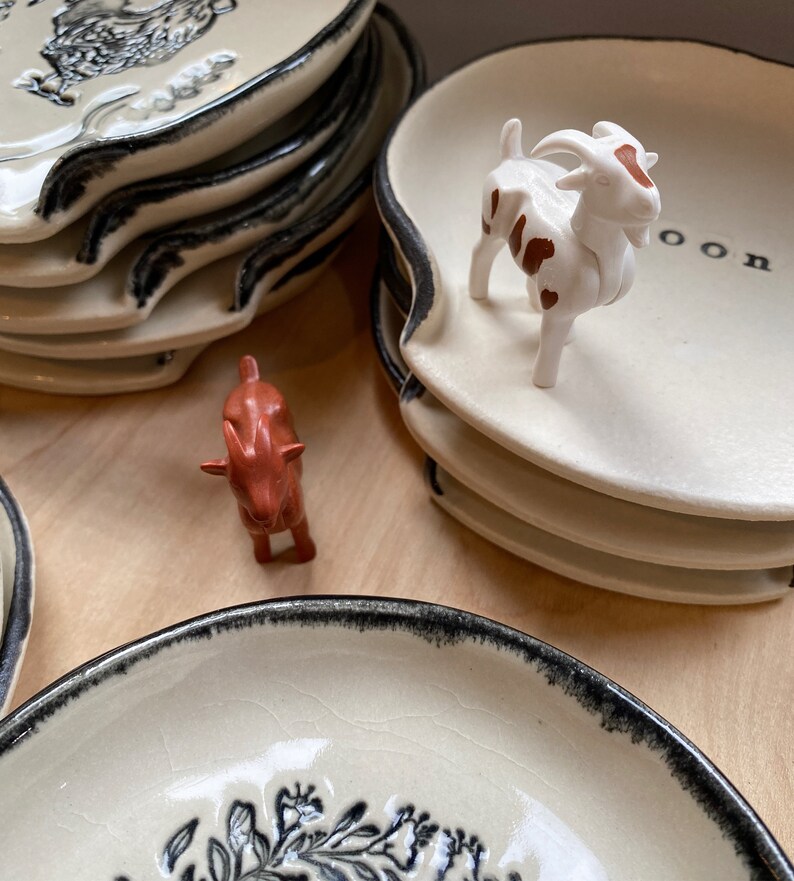 Op de afbeelding: Een stapel cr&egrave;mekleurige keramische lepelsteunen met een zwarte rand en een decoratief ontwerp. Een klein wit geitenbeeldje met bruine vlekken staat op een van de lepelsteunen. Er is ook een ander klein, roodbruin dierenbeeldje aanwezig.