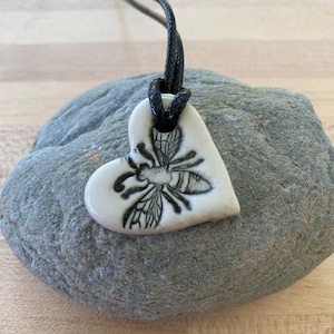 May include: A white ceramic heart-shaped pendant with a black and white bee design hangs from a black cord. The pendant is resting on a gray rock.