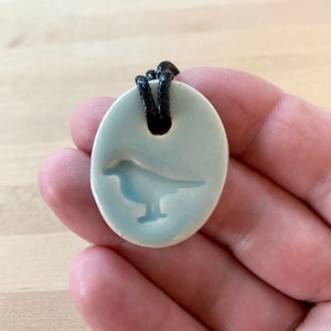 May include: A light blue ceramic pendant with a bird silhouette. The pendant is attached to a black cord.