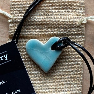 May include: A light blue ceramic heart pendant with a crackled glaze, hanging from a black cord necklace. The necklace is in a small burlap pouch.