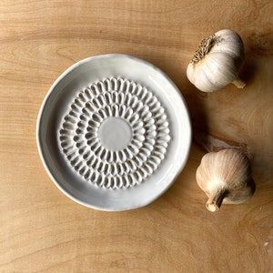 May include: White ceramic garlic press with a textured surface. Two whole garlic bulbs are on a wooden surface.