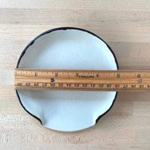 Op de afbeelding: Een kleine, cr&egrave;mekleurige keramische schaal met een zwarte rand en twee kleine handvatten. Een houten liniaal geeft aan dat de schaal ongeveer 15 cm in diameter is. De schaal staat op een licht houten oppervlak.