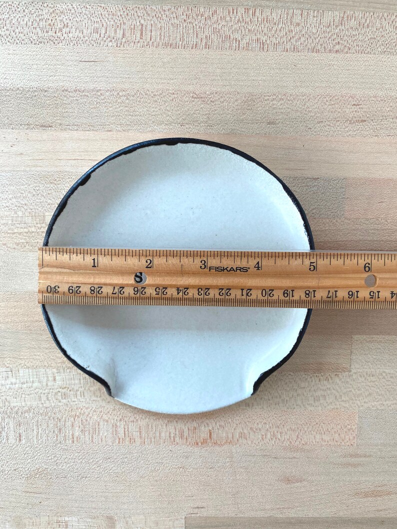 Op de afbeelding: Een kleine, cr&egrave;mekleurige keramische schaal met een zwarte rand en twee kleine handvatten. Een houten liniaal geeft aan dat de schaal ongeveer 15 cm in diameter is. De schaal staat op een licht houten oppervlak.