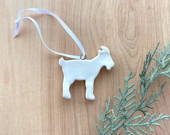 May include: White ceramic goat ornament with a white ribbon for hanging. The ornament is on a light brown wooden surface.