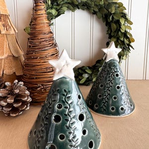 May include: Two ceramic Christmas tree figurines with white star toppers. The trees are dark green with a glossy finish and feature cut-out circles and leaf designs. A woven tree and a green wreath are in the background.