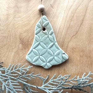 May include: A light blue ceramic bell ornament with a geometric pattern. The bell is hanging from a twine string with a wooden bead.
