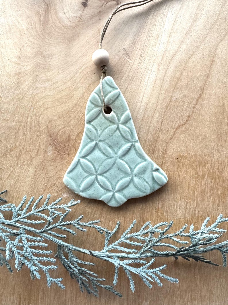 May include: A light blue ceramic bell ornament with a geometric pattern. The bell is hanging from a twine string with a wooden bead.
